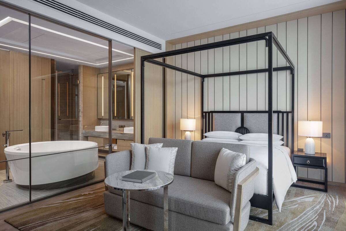 A modern hotel room with a four-poster bed, a sitting area with a small table, a bathtub, and a large mirror.