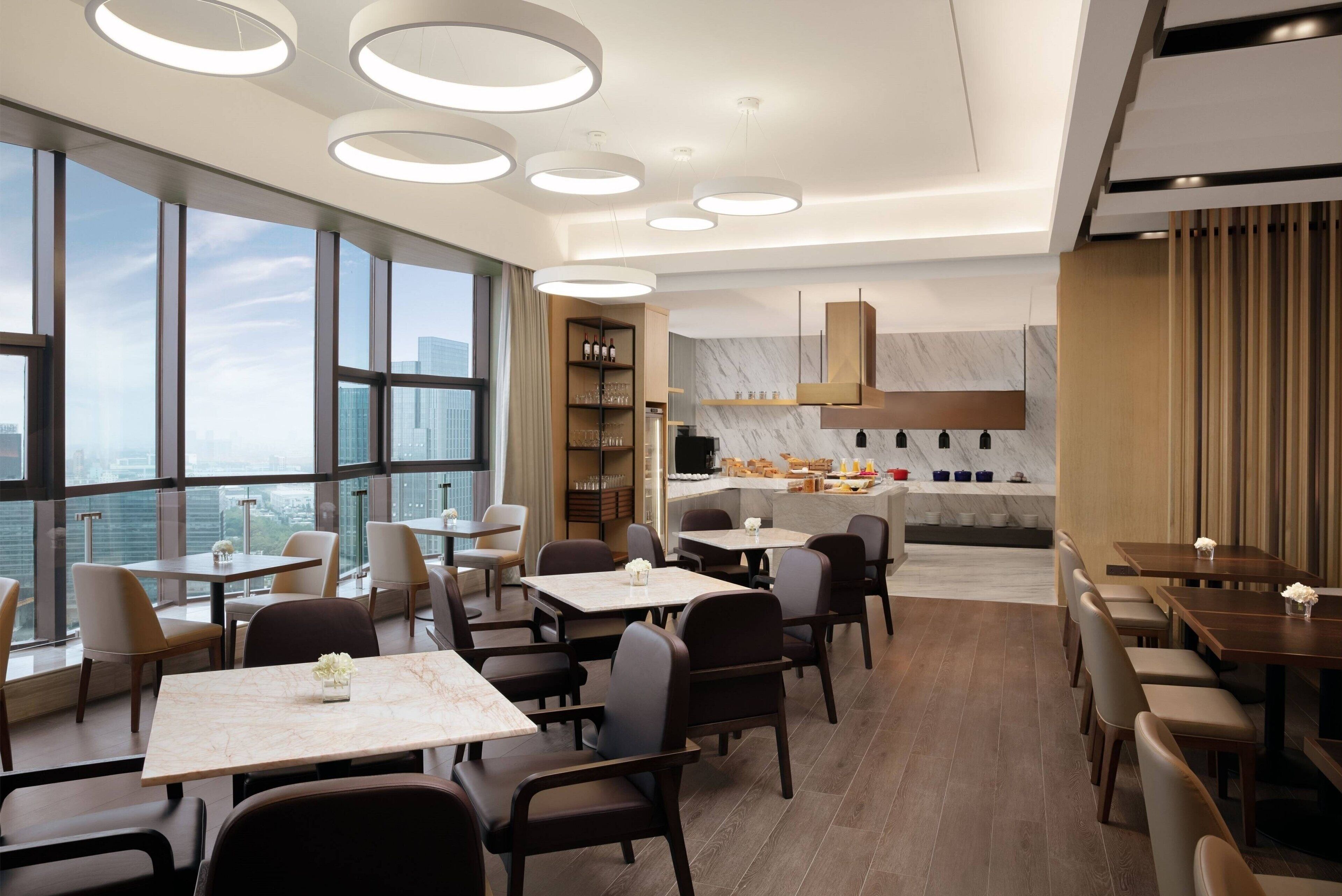 A modern dining area with round tables and chairs, a kitchen in the background, and large windows offering a city view.