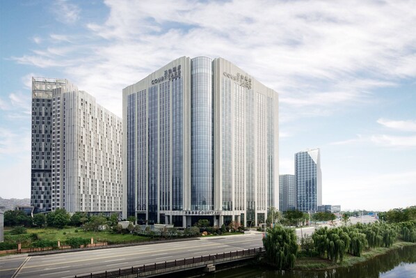 A modern high-rise building with a prominent 'Courtyard' sign, surrounded by greenery and a water body. - Courtyard by Marriott Jiangsu Taizhou (Taizhou)