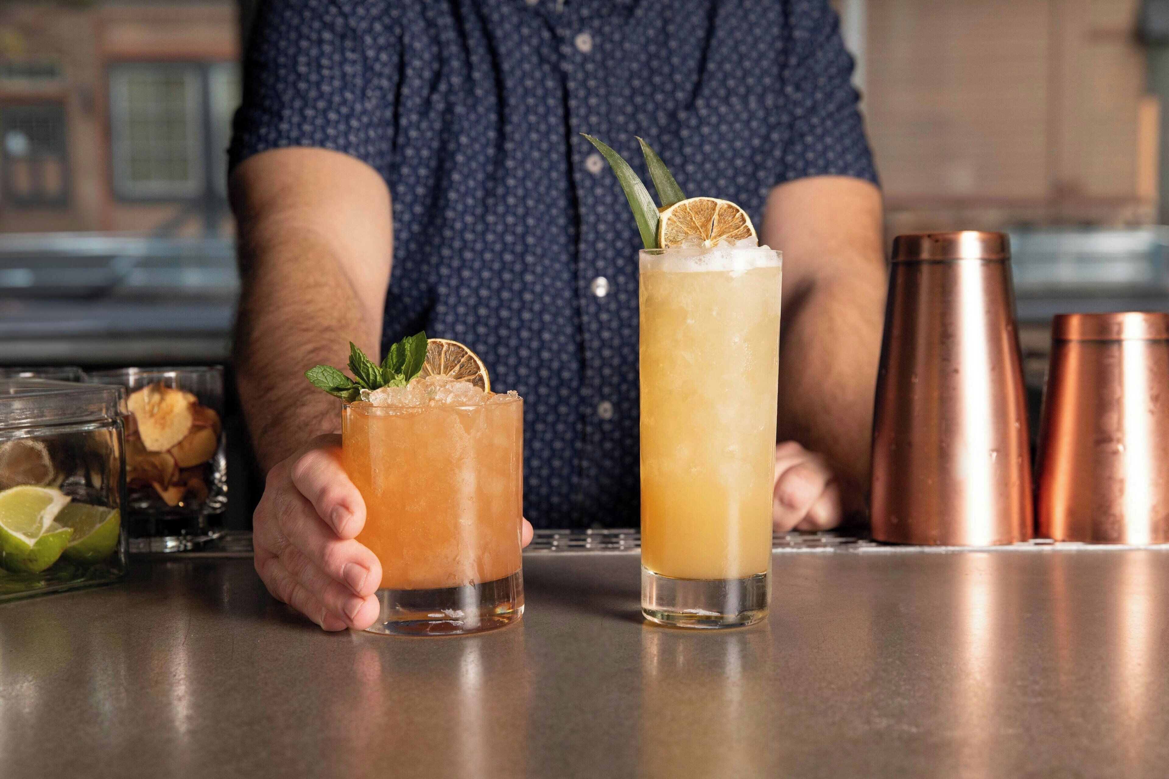 Two cocktails on a bar counter with a person holding one.