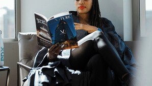 Une personne est assise sur une chaise, lisant un livre dans une pièce qui possède une fenêtre et une lampe.