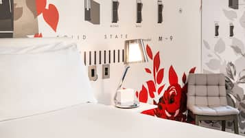 A hotel room with a bed, a bedside table with a lamp, a chair, and a wall with audio equipment labels.