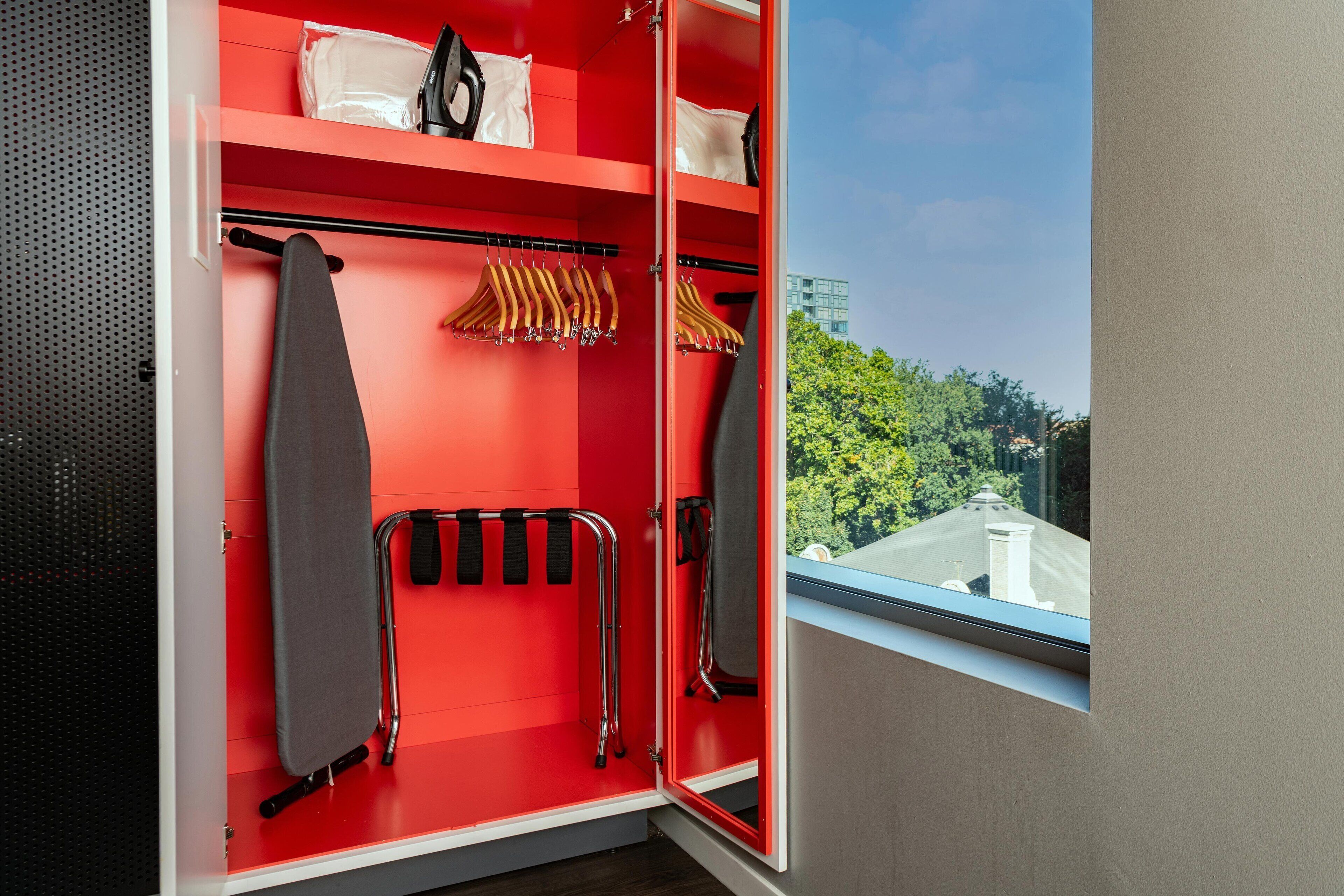 A red wardrobe with hanging space for clothes, a mirror, and a window with a view of trees and buildings.