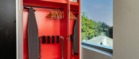 A red wardrobe with hanging space for clothes, a mirror, and a window with a view of trees and buildings.