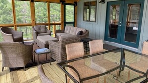 Un'area salotto esterna coperta con mobili in vimini e un tavolo in vetro, con vista su una zona boschiva.