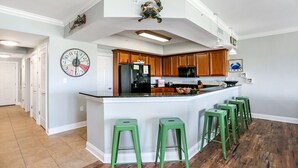 A modern kitchen with a central island, green bar stools, wooden cabinets, and a decorative clock. - Gulf View Condo~ Paradise Shores 403 Sunset Paradise (Mexico Beach)