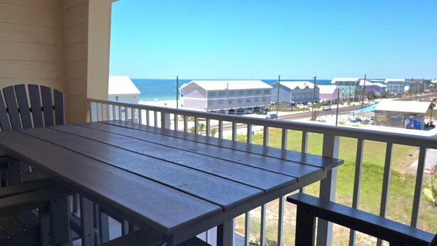 A balcony with a wooden table and chairs, overlooking a coastal area with buildings and a clear sky. - Gulf View Condo~ Paradise Shores 403 Sunset Paradise (Mexico Beach)