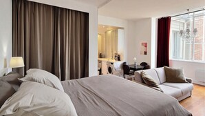 Ein Hotelzimmer mit einem großen Bett, einer Couch, einem Schreibtisch mit Stuhl, einer Waschmaschine und einem großen Fenster mit Vorhängen.