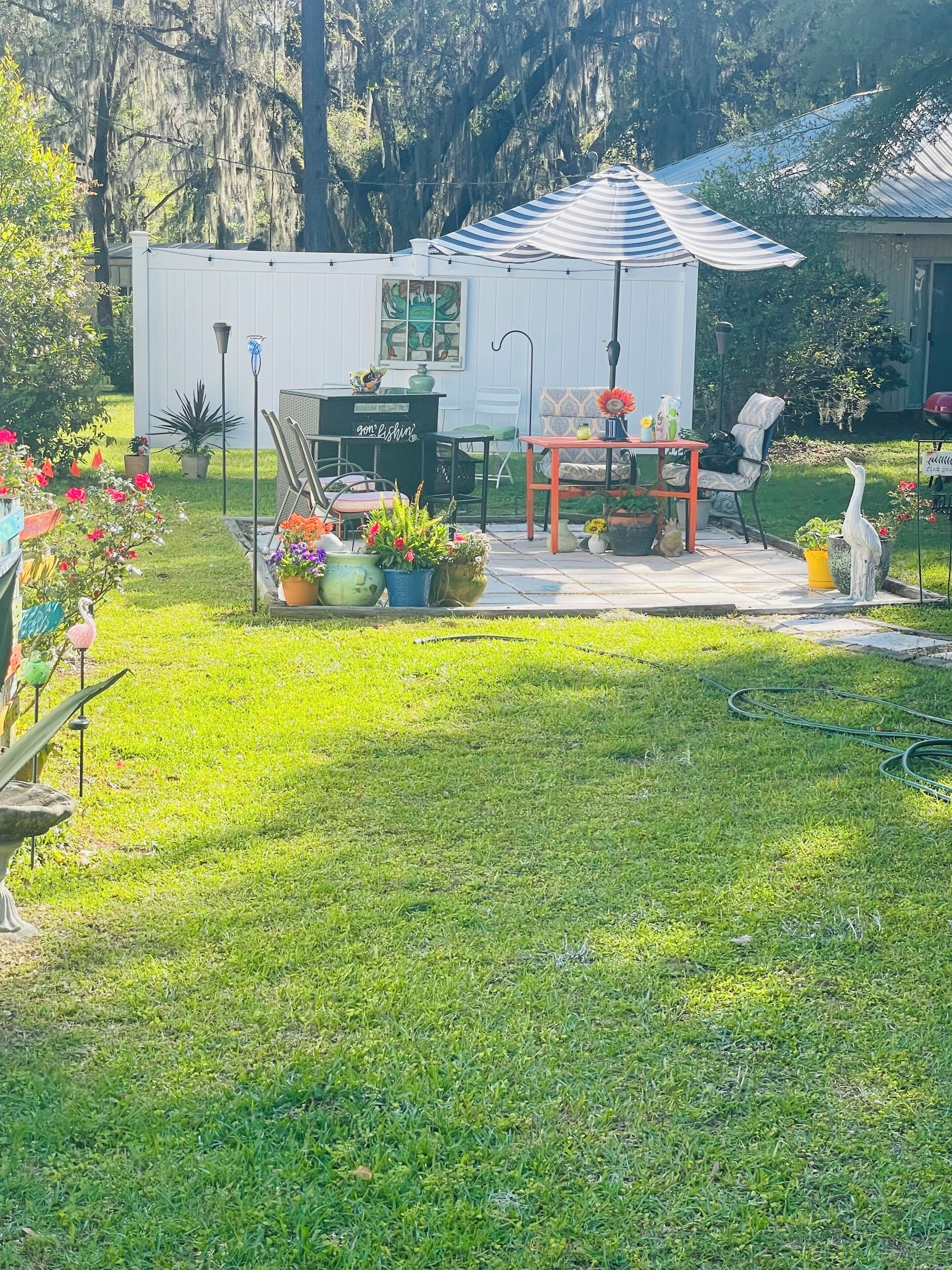 Una casa pequeña y blanca con un porche, una mesa de comedor y sillas. Hay plantas en macetas y una estatua de pájaro en el césped.