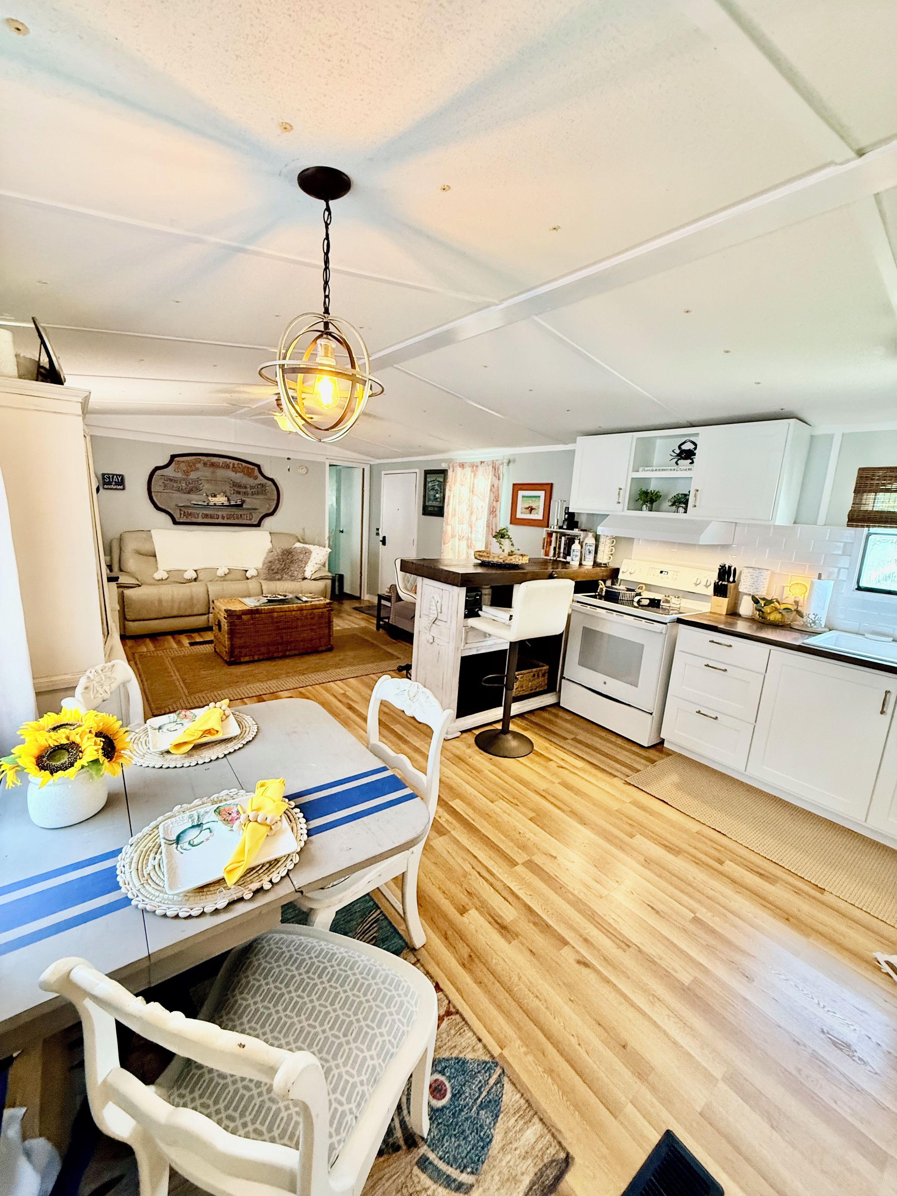 A spacious kitchen with white cabinetry, a dining area with a table set for two, and a living room with a sofa and a decorative wall.