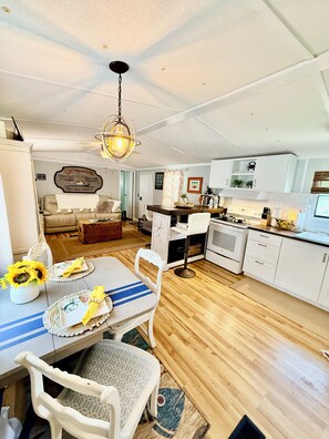 A spacious kitchen with white cabinetry, a dining area with a table set for two, and a living room with a sofa and a decorative wall.