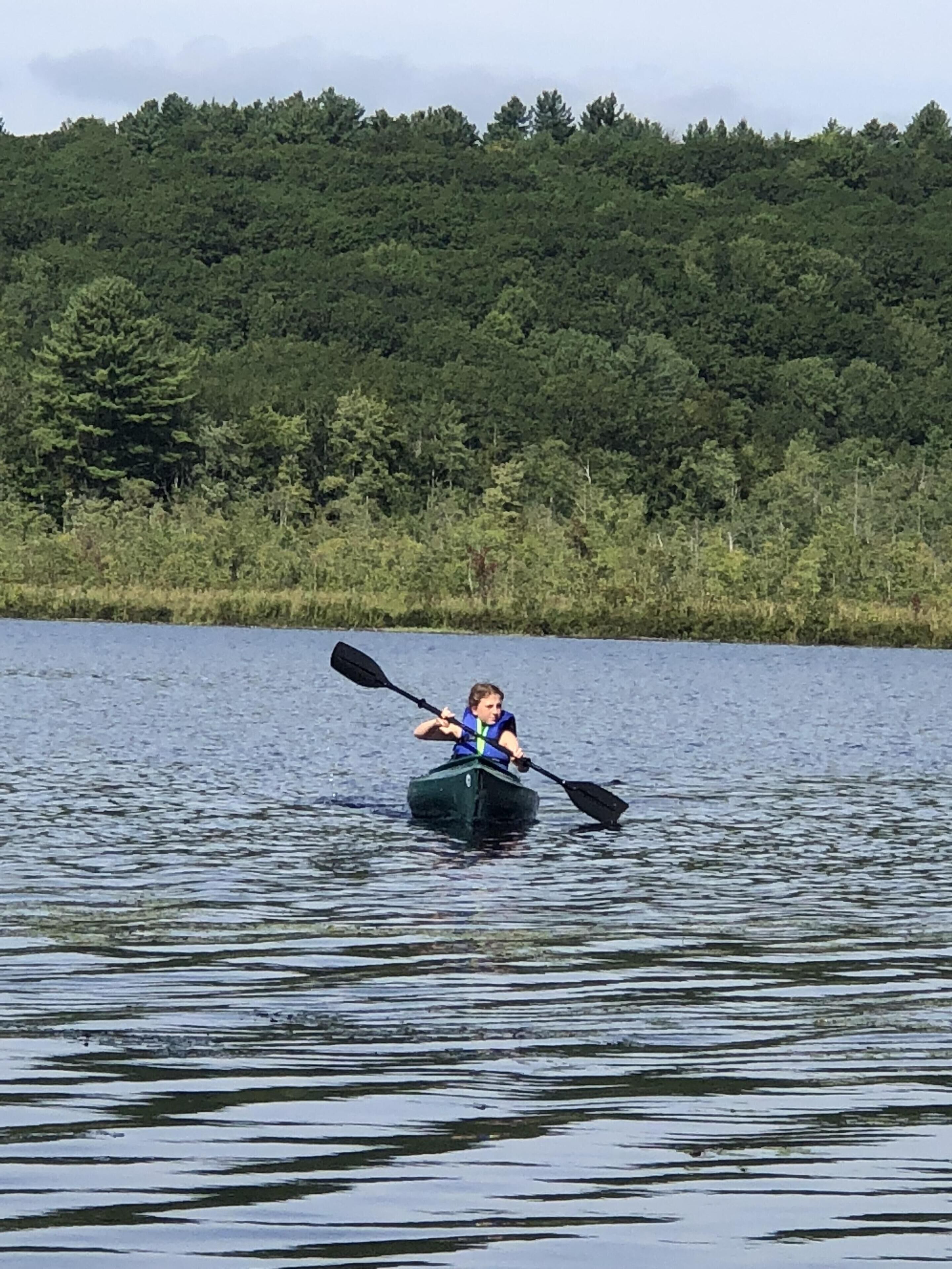 Une personne pratiquant le kayak sur un lac paisible, entouré d’une forêt dense.
