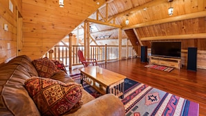 A spacious loft with a high ceiling, wooden beams, a large TV, a sofa, and a red chair. - Luxurious Woodstock Home Rental Catskill Mountains Views & Private Retreat (Bearsville)