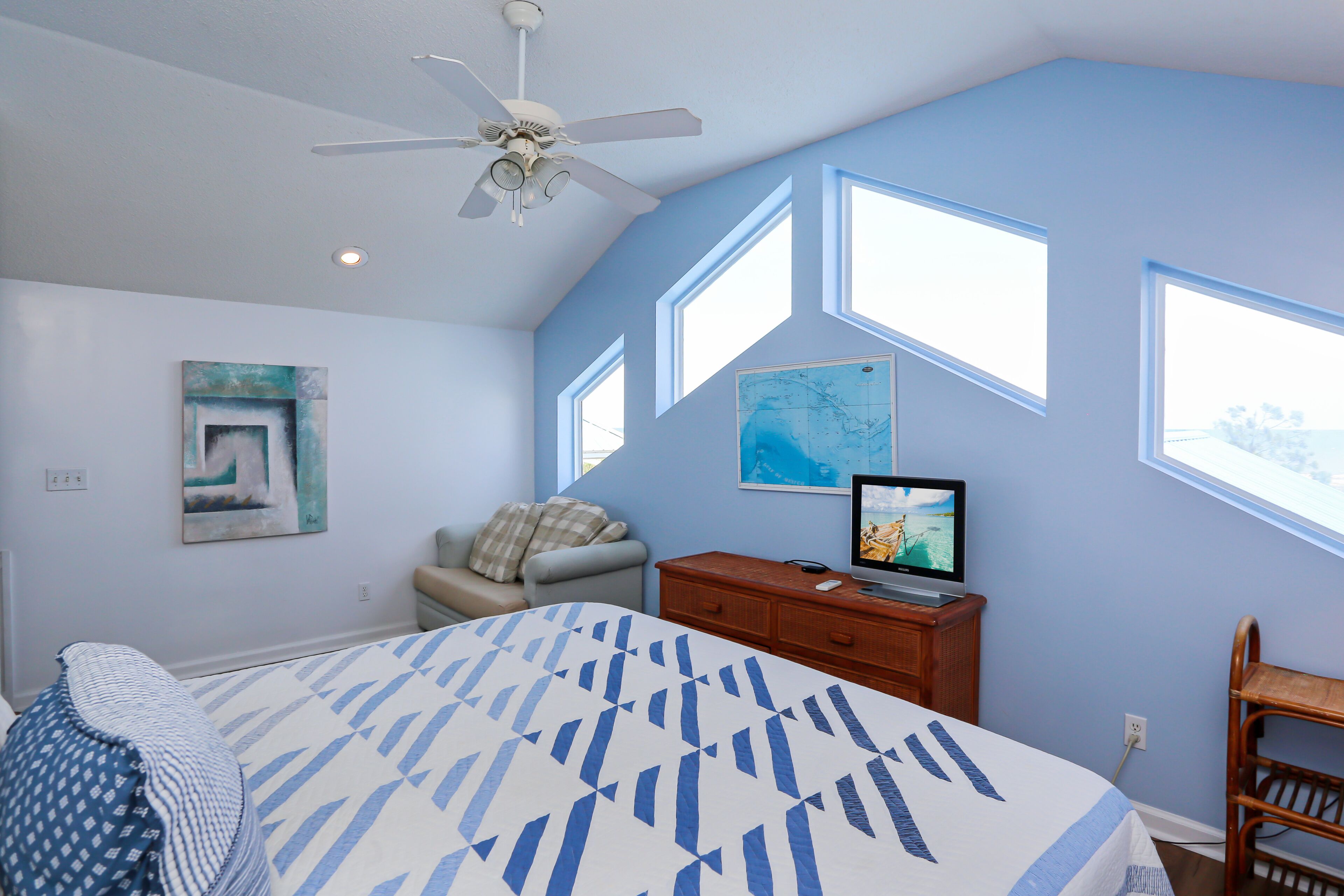 A bedroom with a bed, a desk, a chair, a ceiling fan, and large windows.