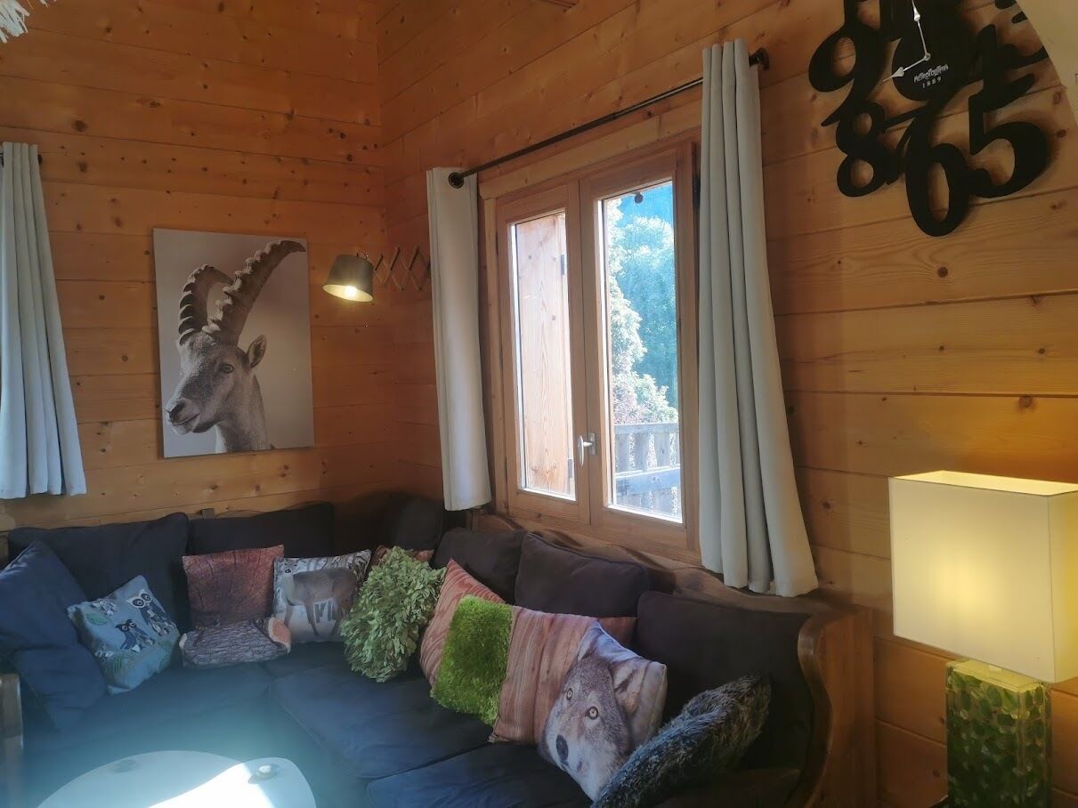 A cozy cabin interior with a wooden wall, a sofa with animal-themed cushions, a framed animal picture, and a window with curtains.