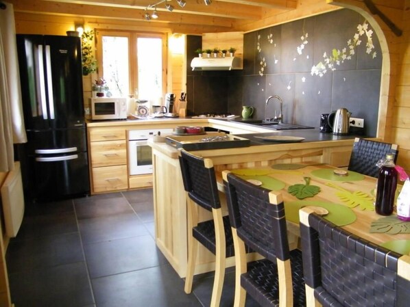 Cozinha moderna com armários de madeira, geladeira preta, micro-ondas e pia. Há uma mesa de jantar com cadeiras e um painel decorativo.