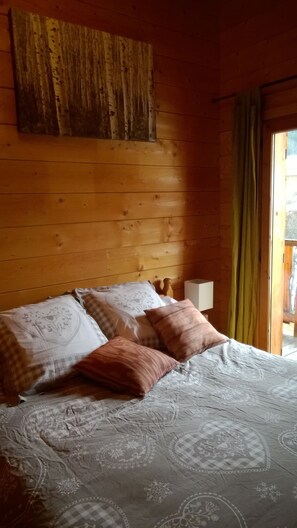 Quarto de cabana de madeira com cama, almofadas, mesa de cabeceira e janela com cortinas.