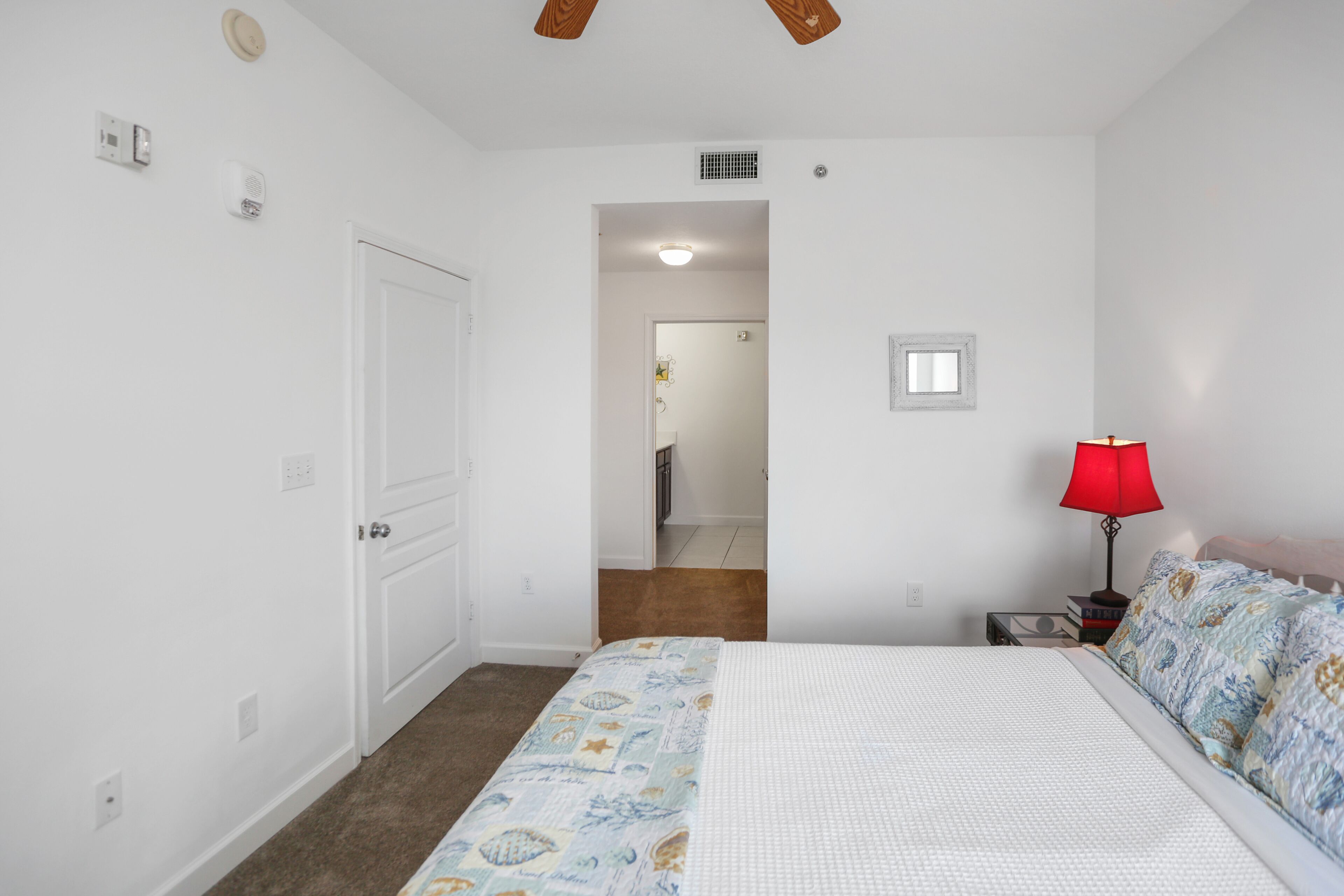 A bedroom with a bed, a nightstand, a red lamp, and a door leading to another room.
