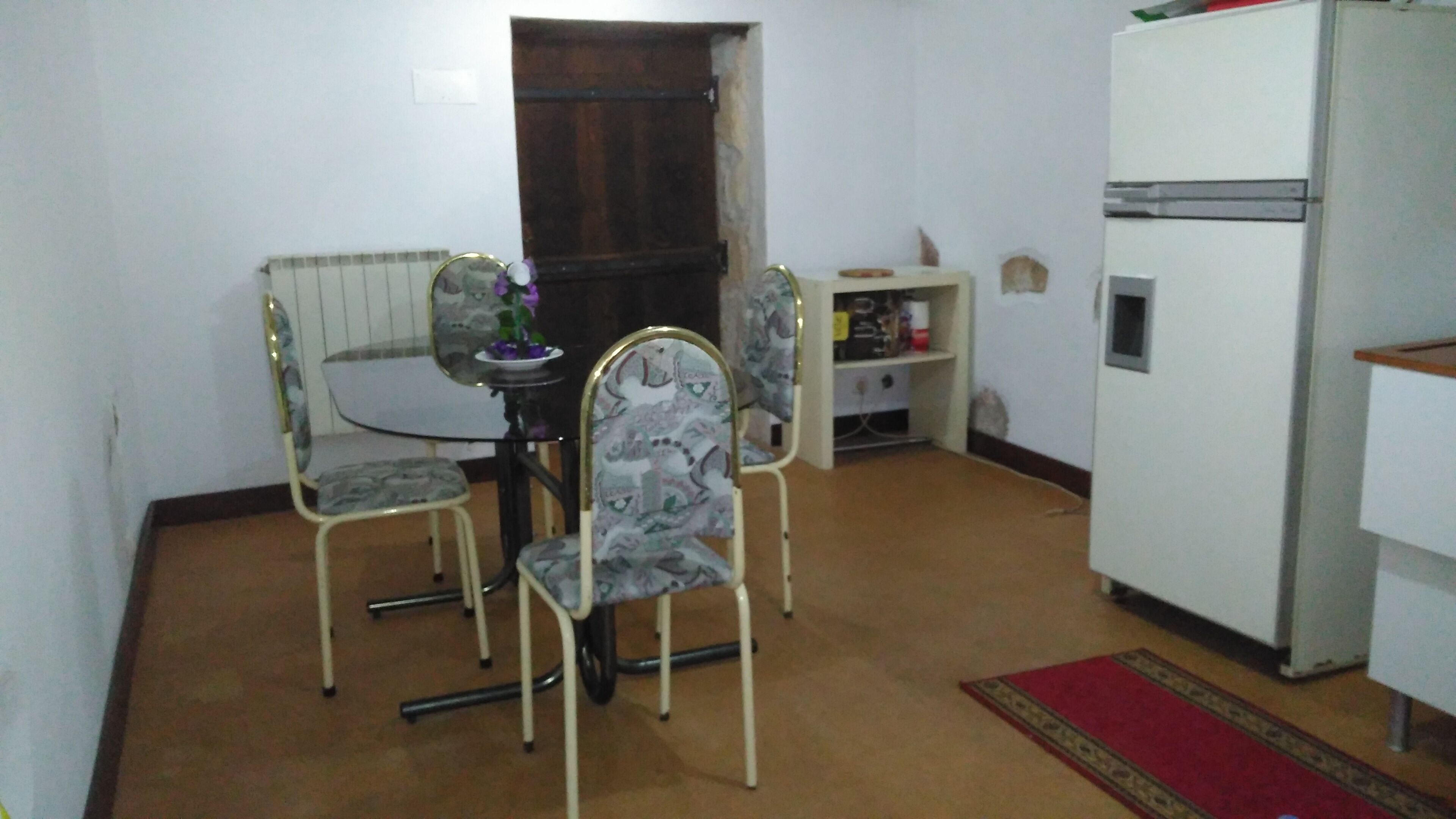 A small, compact room with a dining table and four chairs, a refrigerator, and a radiator.