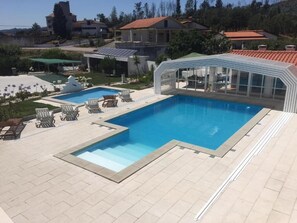 A swimming pool with lounge chairs and a glass-enclosed area, surrounded by a landscaped garden and residential buildings. - Beautiful family house with pool in the center of a typical village of Portugal (Vila Nova de Poiares)