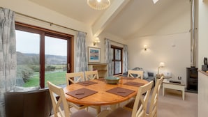Un coin repas avec une table et des chaises en bois, une vue sur la campagne grâce à de grandes fenêtres et une cheminée.