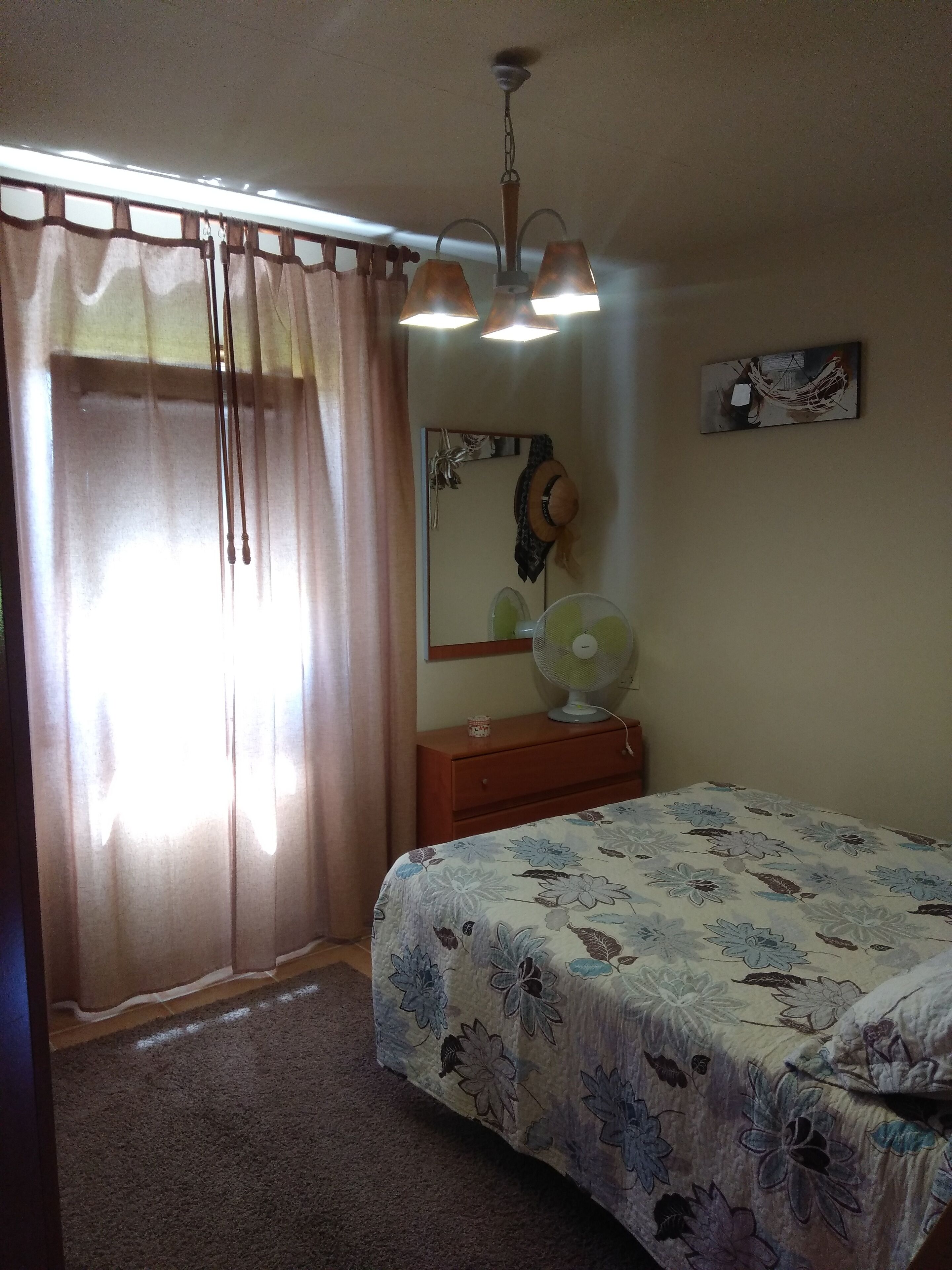 Um quarto com uma cama, mesa de cabeceira, ventilador, espelho e cortina.