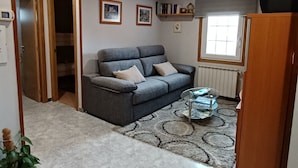 A living room with a grey sofa, a coffee table, and a patterned rug. There is a door leading to another room, a window with blinds, and a small shelf with decorative items. - "EIRA VELLA" Holiday house, (from € 30 day / month). (Pontevedra)