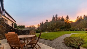 Un patio con una mesa redonda y sillas, un muro de piedra y vistas a un césped con árboles y un atardecer.