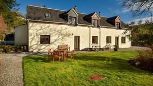 Une maison à deux étages avec un toit en pente, un petit porche à l’avant avec une table et des chaises, et une pelouse bien entretenue.