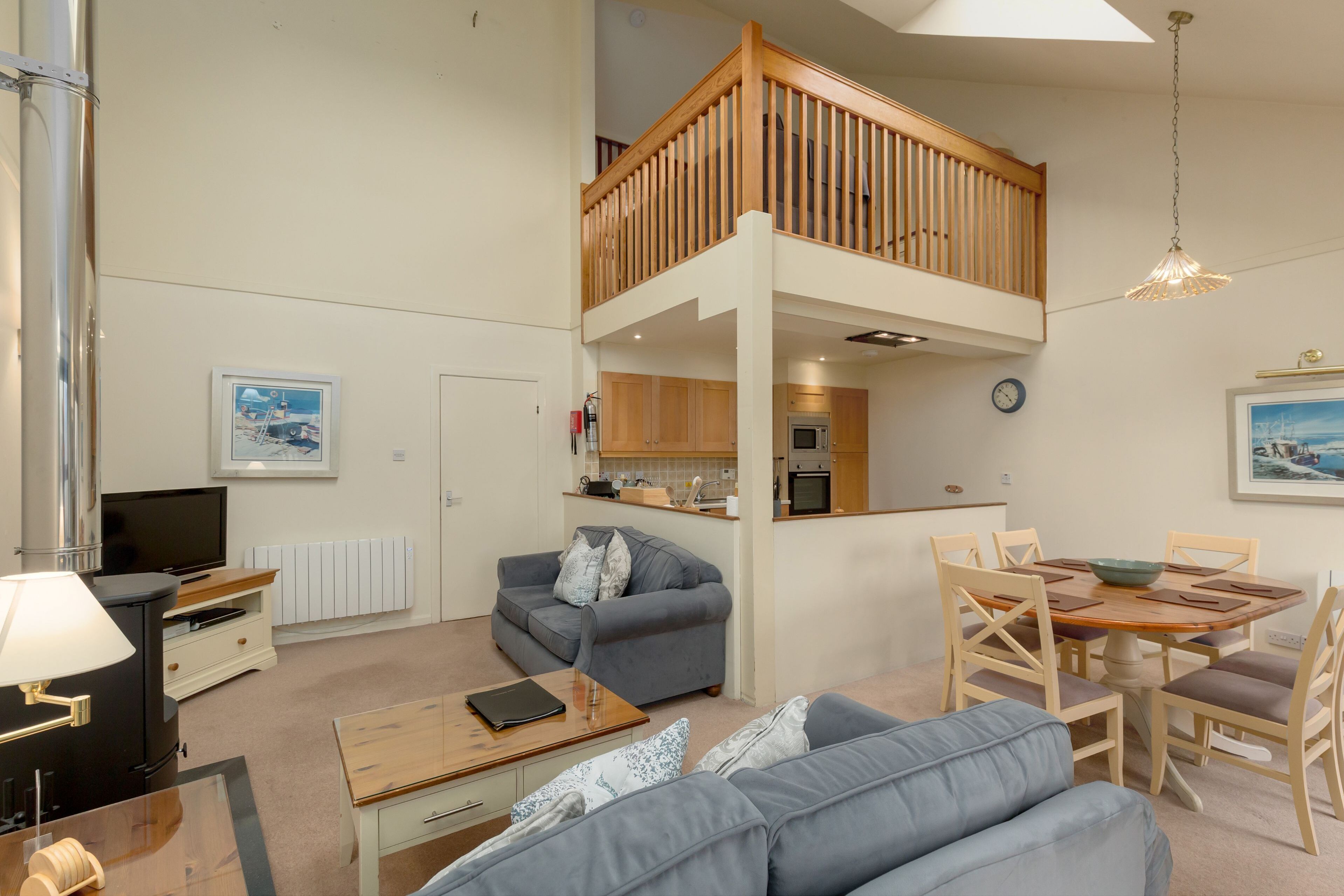 A spacious living area with a high ceiling, a wooden balcony, a kitchen, and a dining table.