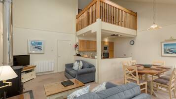 A spacious living area with a high ceiling, a wooden balcony, a kitchen, and a dining table.