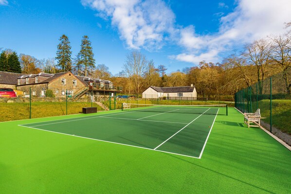 A tennis court with a net, surrounded by a green fence, and adjacent to a building with a stone facade.