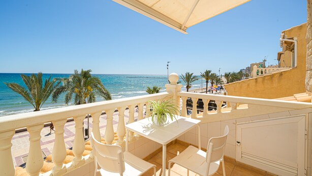 Ein Balkon mit weißem Tisch und Stühlen, Blick auf einen Strand mit Palmen und das Meer.