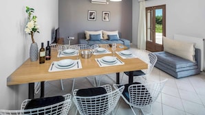A dining area with a wooden table set for four, white wire chairs, a sofa, and a television in the background. - Villa Orchard (Zakynthos)
