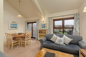 A living room with a grey sofa, wooden dining table, and chairs. There are two windows with curtains, a wall-mounted shelf, and a hanging light fixture. - Walled Garden Mews 5 - a mews cottage that sleeps 6 guests in 2 bedrooms (Oban)