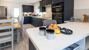 A modern kitchen with dark cabinets, a dining area with a wooden table and chairs, and a breakfast setup with croissants and mugs. - Pier House Two -  Loch-side semi-detached house sleeping up to 10 (Oban)