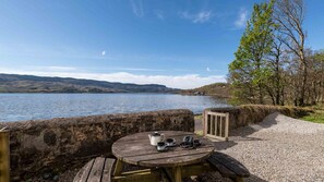 A wooden table with a kettle, cups, and a camera on a gravel path beside a lake with a stone wall and trees.