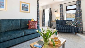 A living room with a blue sofa, a striped armchair, a coffee table with a book and a vase of flowers, and a view of the sea through large windows. - Pier House Two -  Loch-side semi-detached house sleeping up to 10 (Oban)