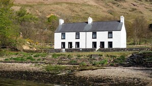 A white building with a grey roof, situated by a body of water with a rocky shoreline and vegetation. - Pier House Two -  Loch-side semi-detached house sleeping up to 10 (Oban)