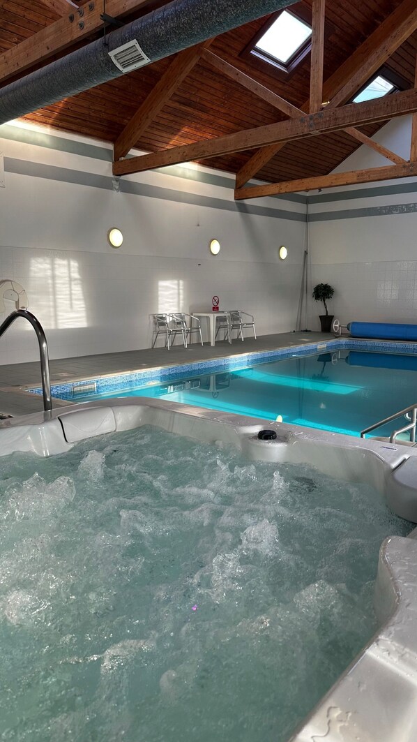 An indoor swimming pool with a hot tub, wooden beams, and a seating area with chairs.
