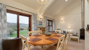 A dining area with a wooden table and chairs, a view of the countryside through large windows, and a fireplace.