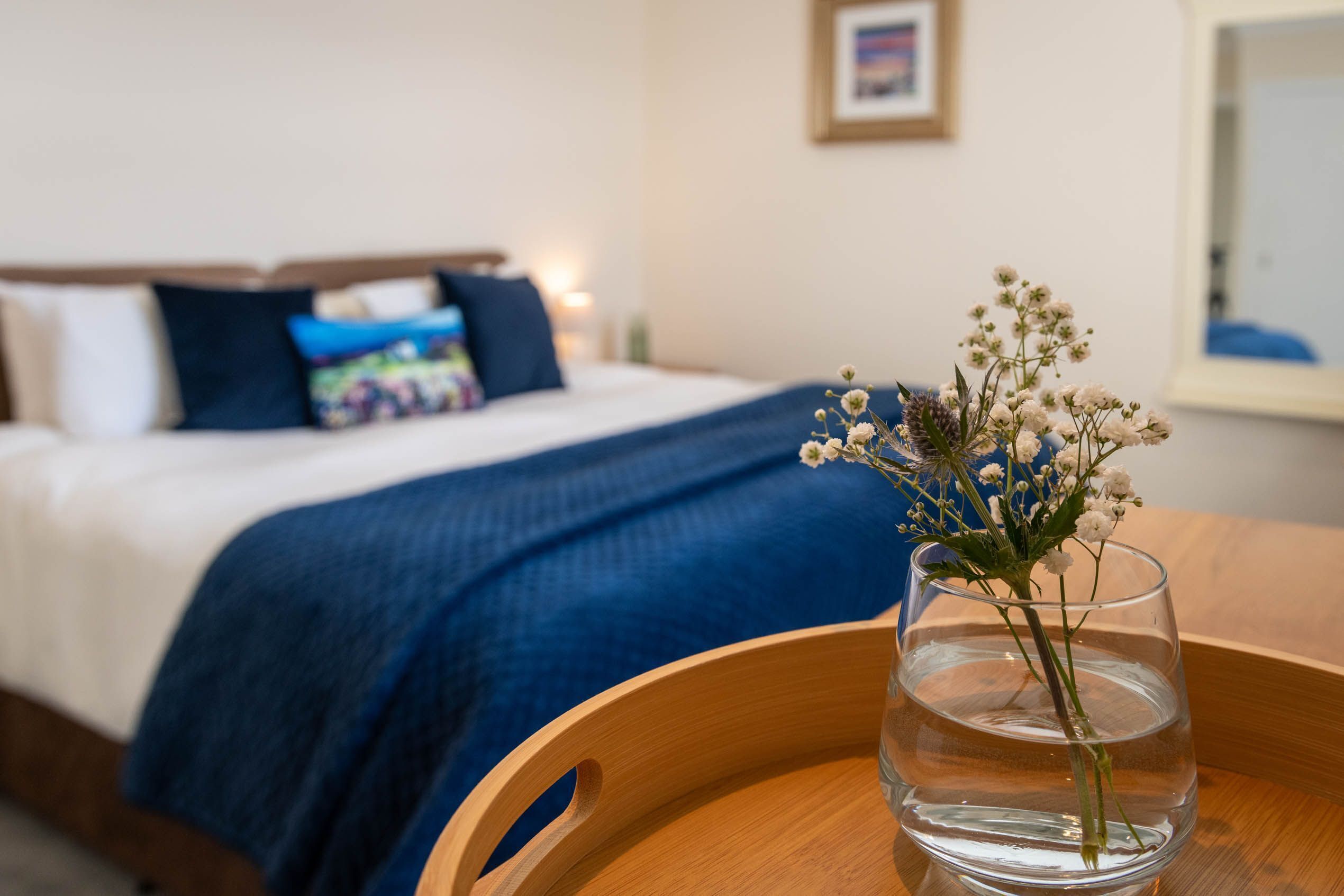 A hotel room with a bed, a blue blanket, a wooden chair, and a vase with flowers.