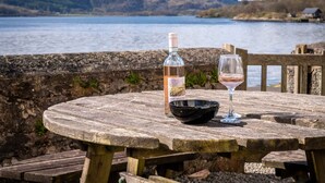A wooden table with a bottle and a glass, set against a scenic lake view. - Pier House 1 - Semi-detached, stone-built cottage on lochside (Oban)