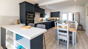 A modern kitchen with dark cabinetry, a central island, and a dining area with a wooden table and chairs. - Pier House 1 - Semi-detached, stone-built cottage on lochside (Oban)