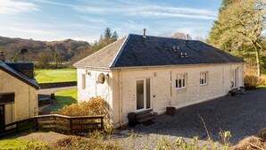 A small, single-story house with a sloped roof, a front door, and a small porch area. - Stables East -  a semi-detached cottage that sleeps up to 6 guests (Oban)