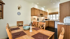 A modern kitchen with wooden cabinets, a dining table with placemats, and a clock on the wall. - Stables East -  a semi-detached cottage that sleeps up to 6 guests (Oban)