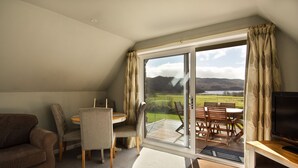 Una sala de estar acogedora con un sofá, una mesa de comedor con sillas, un mueble para la televisión y un amplio ventanal corredizo que da a un patio exterior con vistas panorámicas de colinas y un lago.