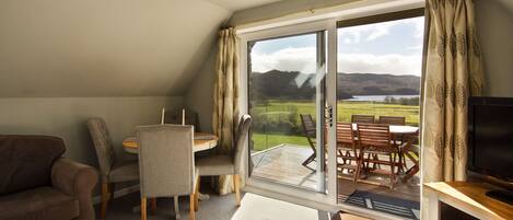 Una sala de estar acogedora con un sofá, una mesa de comedor con sillas, un mueble para la televisión y un amplio ventanal corredizo que da a un patio exterior con vistas panorámicas de colinas y un lago.