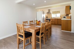 A dining area with a wooden table and chairs, a fruit bowl, and a kitchen in the background.