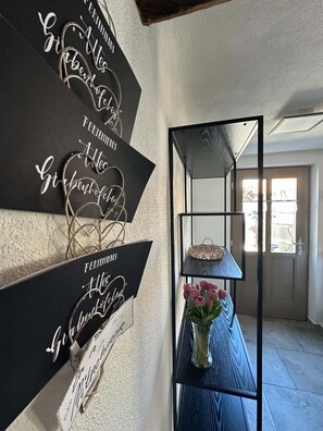 Um corredor com uma placa preta com texto cursivo branco, uma estante de metal com um vaso de flores e uma porta que dá para área externa.